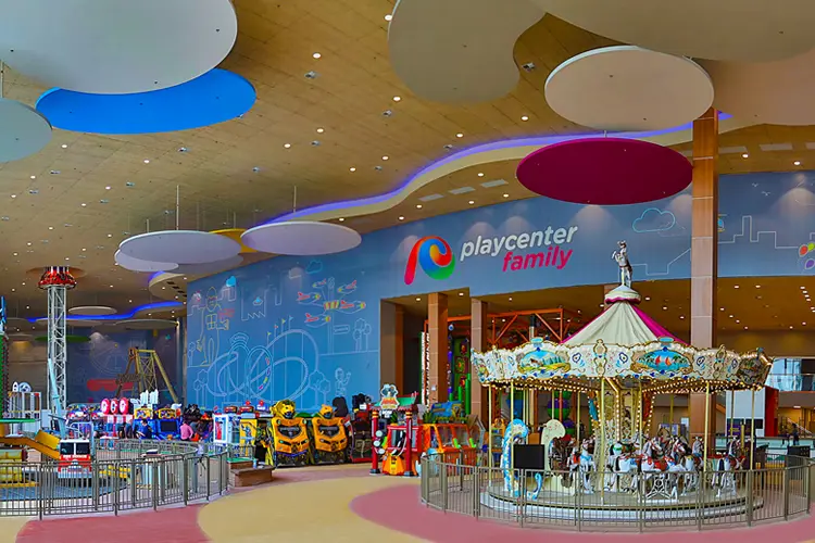 Parque de diversões coberto no Shopping Aricanduva com um carrossel colorido, máquinas de fliperama e um grande muro com o logotipo da Playcenter Family. O teto é adornado com vibrantes decorações circulares penduradas.