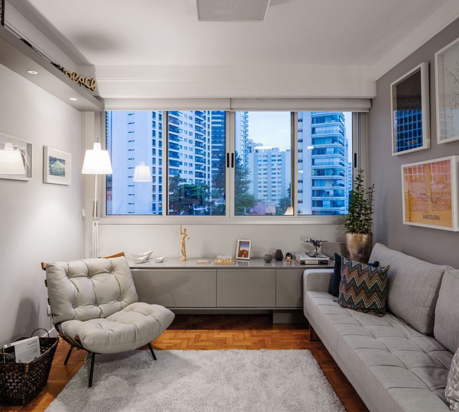Uma sala de estar moderna projetada para decoração de sala pequena apresenta um sofá cinza claro, poltrona acolchoada, aparador baixo, itens decorativos, quadros emoldurados, uma grande janela com vista para a cidade e um tapete cinza no piso de madeira.