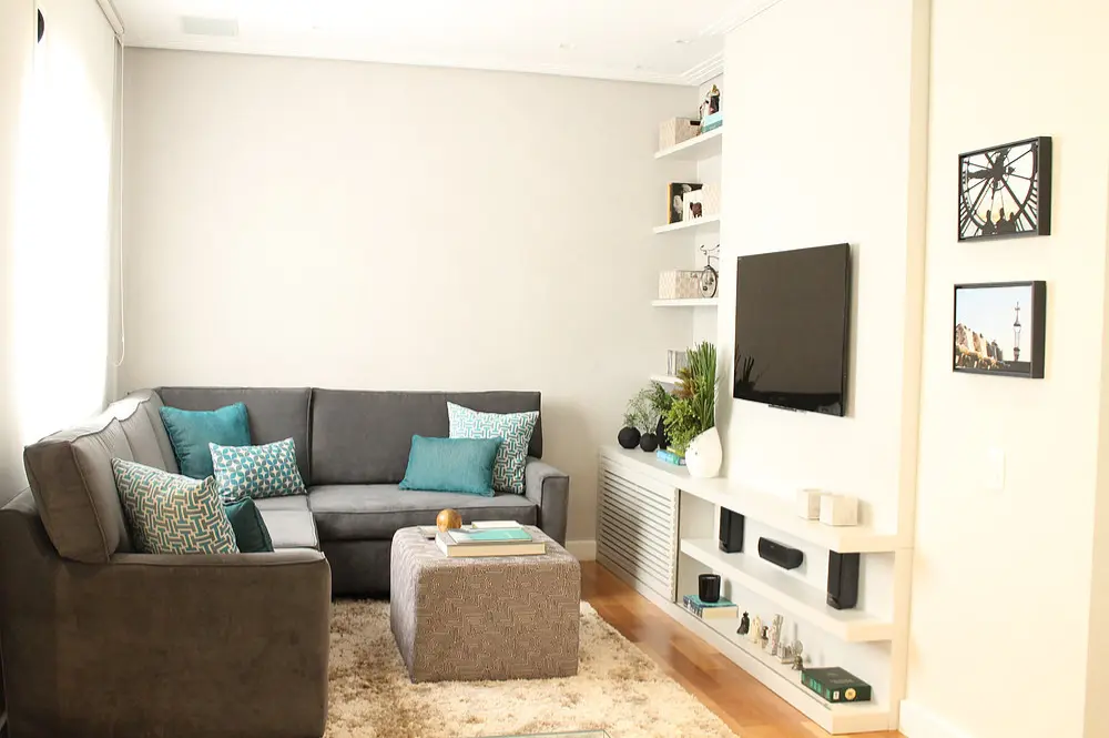 Uma sala de estar moderna com um sofá secional cinza, almofadas azul-petróleo e estampadas, um pufe quadrado, tapete bege, TV na parede, prateleiras embutidas com decoração de sala pequena, paredes de cores claras e piso de madeira.