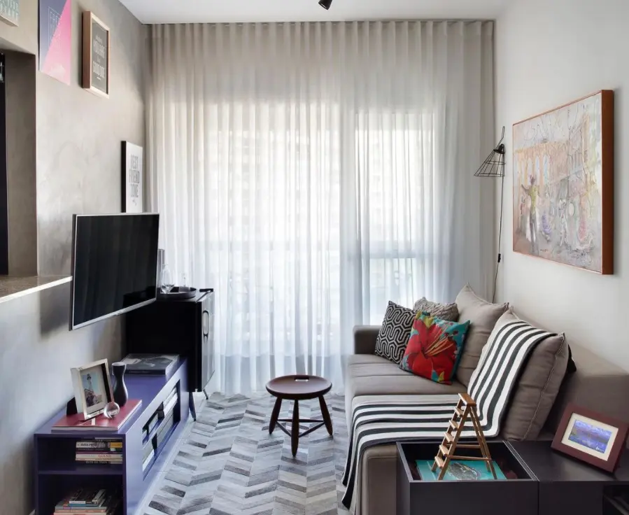 Uma sala de estar moderna com um sofá cinza, almofadas coloridas e decoração de sala pequena, com uma TV montada na parede, estantes de livros, uma pequena mesa de centro redonda e cortinas brancas transparentes cobrindo uma grande janela que deixa entrar luz natural.