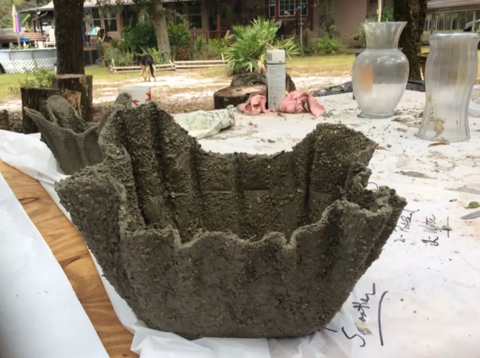 Um vaso de cimento quadrado, de textura áspera e bordas onduladas fica sobre uma superfície de trabalho ao ar livre, cercado por vasos, toalhas e árvores, com casas e um cachorro visíveis ao fundo.