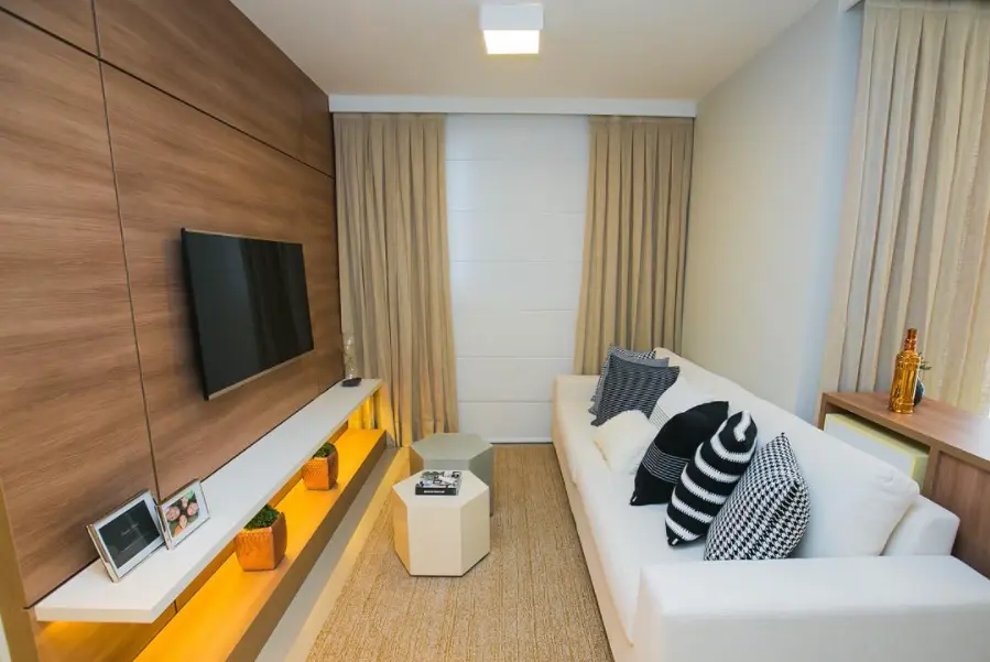 Uma sala de estar moderna com um sofá branco, almofadas estampadas, painéis de madeira e uma prateleira flutuante com fotos emolduradas e plantas exibe uma decoração perfeita para sala pequena, completa com cortinas neutras e uma pequena mesa de centro hexagonal.