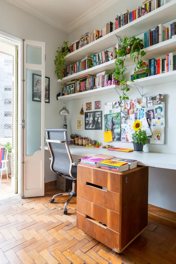 Um home office iluminado no Apartamento Higienópolis conta com uma mesa de madeira, cadeira ergonômica e prateleiras repletas de livros e plantas. Um quadro de cortiça com fotos e anotações fica acima da mesa, enquanto a luz do sol entra por uma porta de vidro para a varanda.