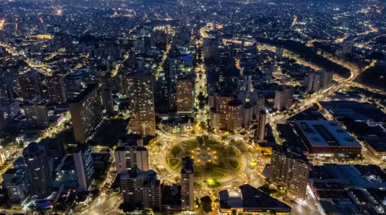 Vista aérea de uma grande cidade à noite, com prédios altos e postes de luz brilhantes, destacando-se o impressionante grafite em BH, próximo a um parque circular no centro. Ruas iluminadas criam um cenário urbano vibrante e animado.