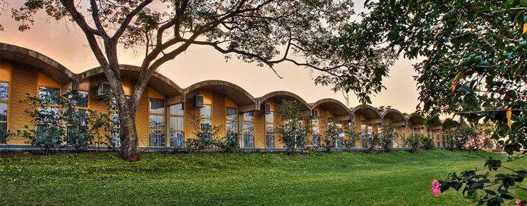 Um edifício moderno com um telhado curvo característico e grandes janelas fica em uma colina gramada, cercado por árvores e vegetação exuberante durante o pôr do sol, refletindo o design inovador frequentemente visto em faculdades em Ribeirão Preto.