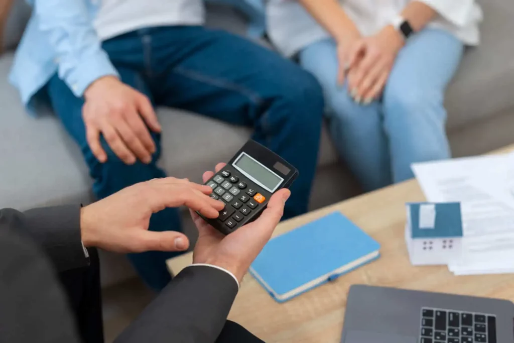Imagem das mãos de um homem fazendo cálculos em uma calculadora ao lado de um casal que está sentado em um sofá em frente a uma mesa de centro onde estão alguns papéis para ilustrar matéria sobre como funciona o feirão de imóveis da Caixa