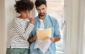 Imagem de um casal formado por um homem e uma mulher encostados em uma janela olhando um documento que está na mão do homem para ilustrar matéria sobre como funciona o direito de preferência
