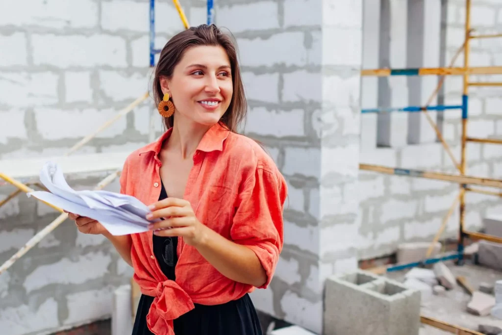 Imagem de uma mulher vestindo uma camisa laranja brilhante segura papéis e sorri enquanto está em um canteiro de obras entre paredes de tijolos inacabadas e andaimes ao fundo