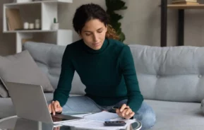 Imagem de uma mulher sentada em um sofá, trabalhando em uma mesa baixa com um laptop, papéis, uma caneta e uma calculadora. Ela parece concentrada, analisando documentos e fazendo anotações sobre o custo efetivo total em um ambiente aconchegante de sala de estar
