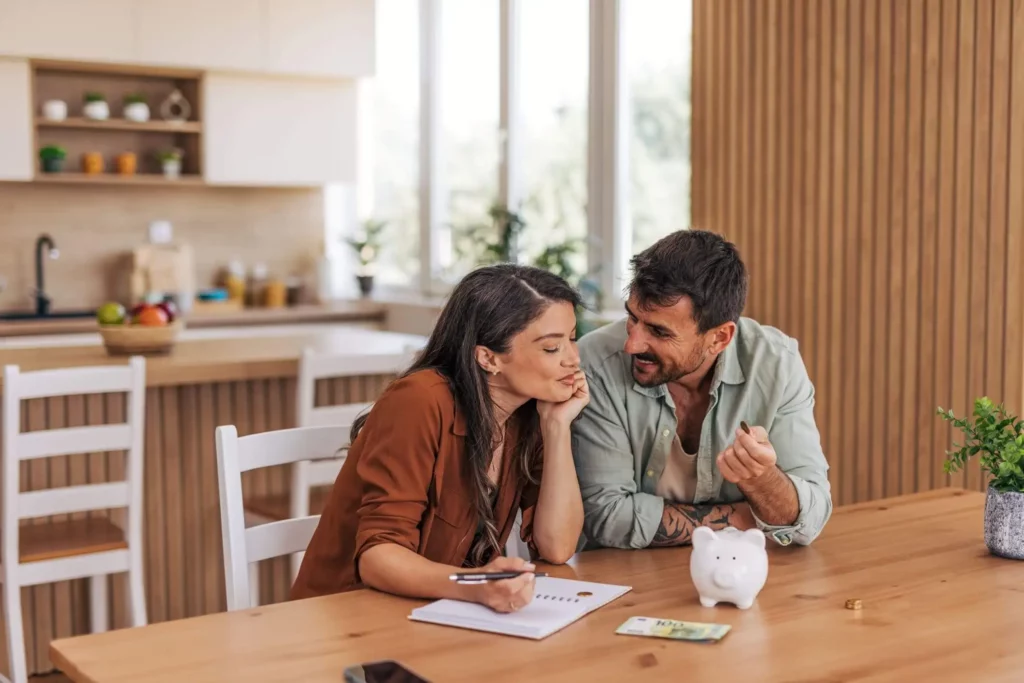 A mulher escreve em um caderno, enquanto o homem gesticula em direção ao cofrinho e ao dinheiro enquanto revisam o custo efetivo total. Uma cozinha moderna pode ser vista ao fundo