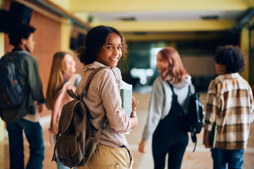 Imagem de uma garota andando com uma mochila nas costas e um caderno na mão enquanto vira o corpo e olha para trás com um sorriso para ilustrar matéria sobre escola em Nilópolis