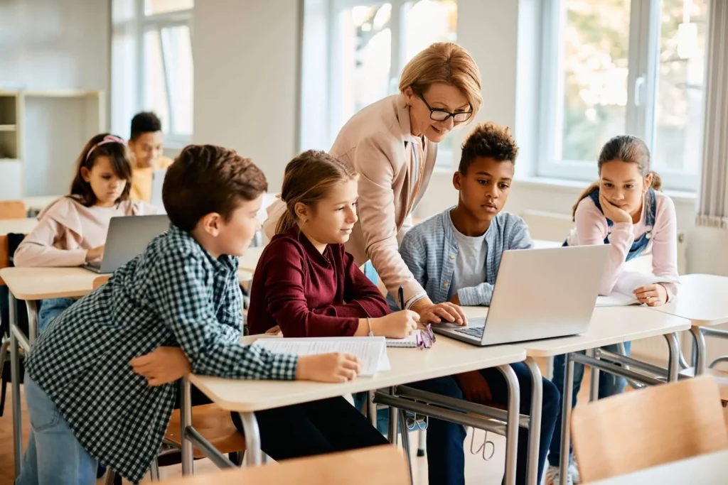 Imagem de uma professora ensinando uma lição para quatro alunos sentados alinhados em carteiras enquantos eles olham para a tela de um computador para ilustrar matéria sobre as escolas em Nilópolis