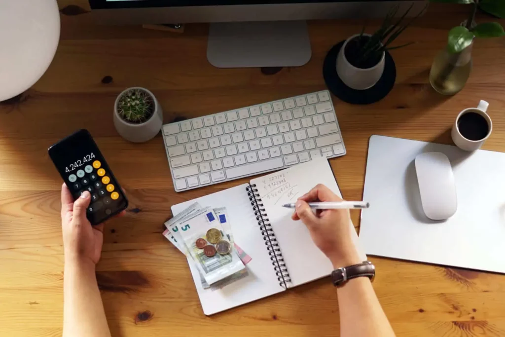 Imagem de uma pessoa calculando despesas usando uma calculadora de telefone, com notas e moedas de euro em um caderno, para ilustrar matéria sobre se tem como comprar casa sem entrada. Na mesa há um teclado, mouse, xícara de café e vasos de plantas.