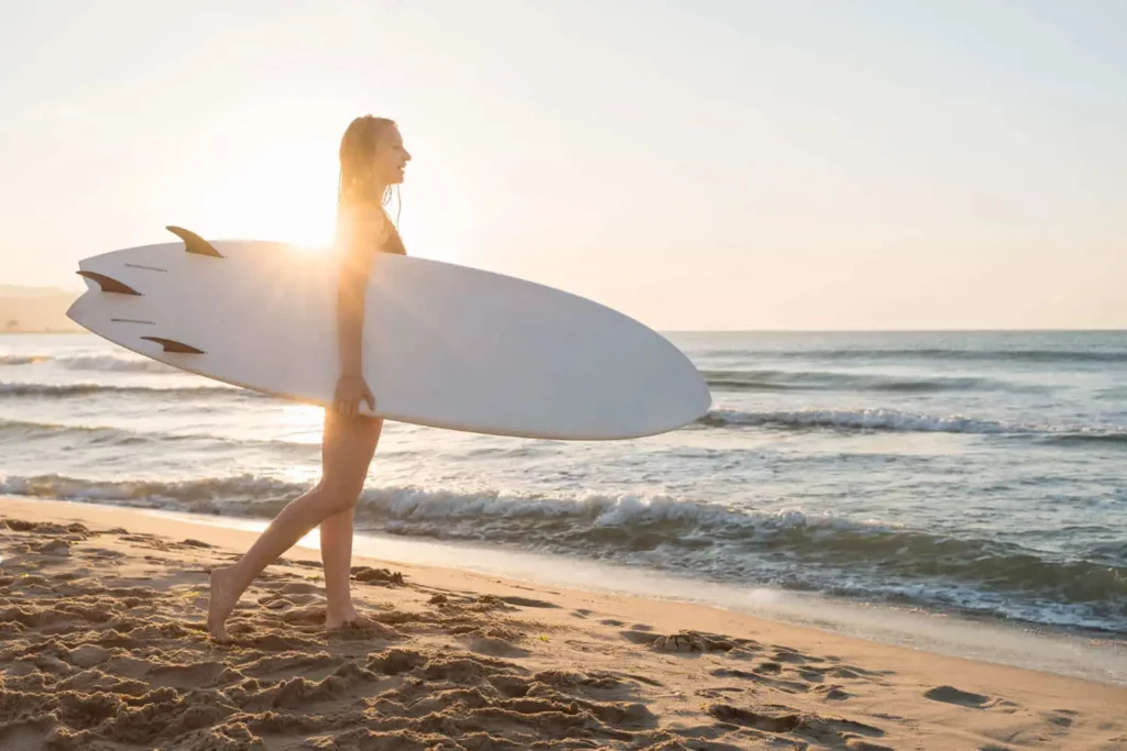Imagem de uma mulher jovem segurando uma prancha de surfe em frente ao mar durante o entardecer para ilustrar matéria sobre o mapa das praias de Ubatuba