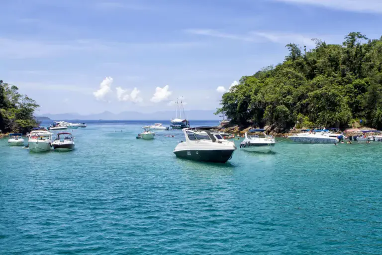 Imagem de barcos ancorados em águas cristalinas azul-turquesa perto de uma ilha exuberante e coberta de árvores sob um céu azul brilhante em Angra dos Reis, no Rio de Janeiro