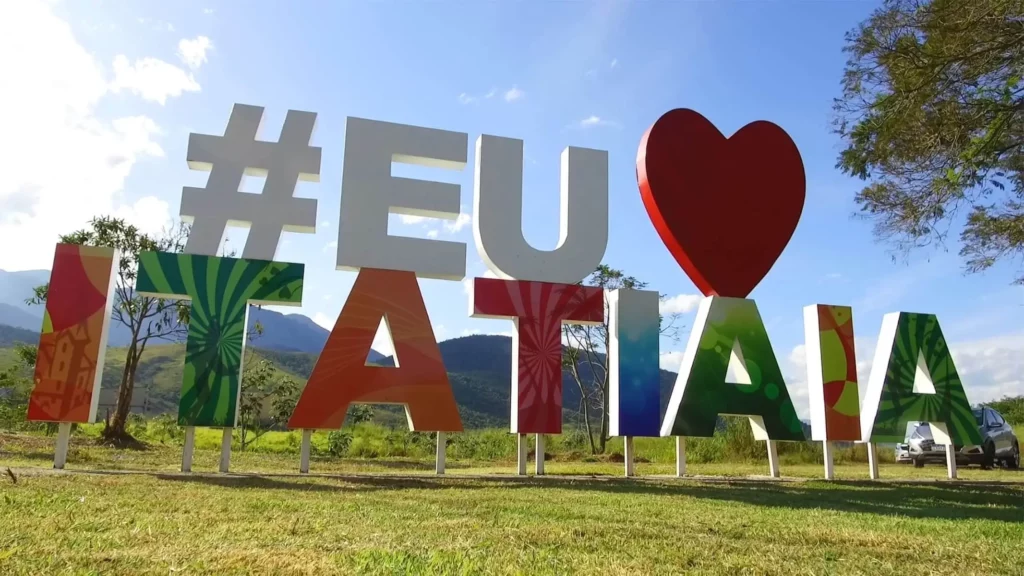 Imagem de um grande letreiro ao ar livre dizendo “Eu amo Itatiaia” com letras coloridas, em meio a colinas verdes, árvores e um céu azul em Itatiaia, no Rio de Janeiro