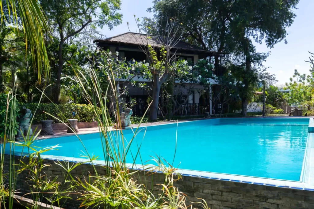 Imagem de uma piscina azul clara cercada por plantas e árvores verdejantes, com uma casa de dois andares ao fundo parcialmente sombreada pela folhagem, ilustra bem a diferença entre chácara ou sítio na vida rural.