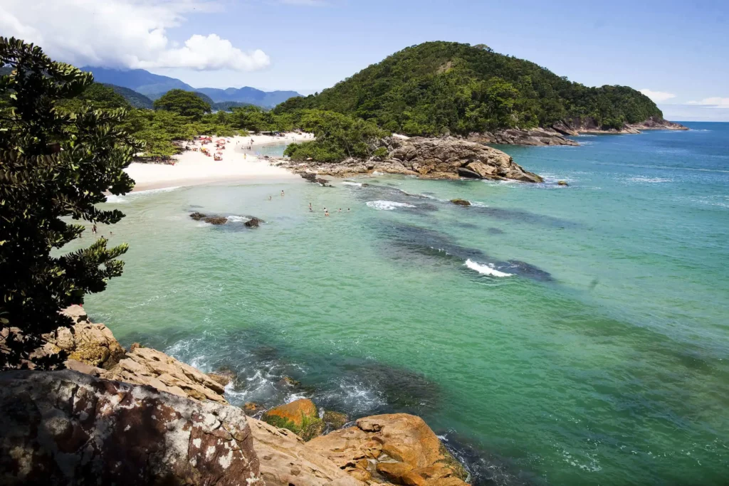 Imagem de uma praia em Trindade, no interior do Rio de Janeiro, mostra água azul-turquesa clara, afloramentos rochosos, colinas verdes e pessoas relaxando na areia sob um céu azul com nuvens dispersas