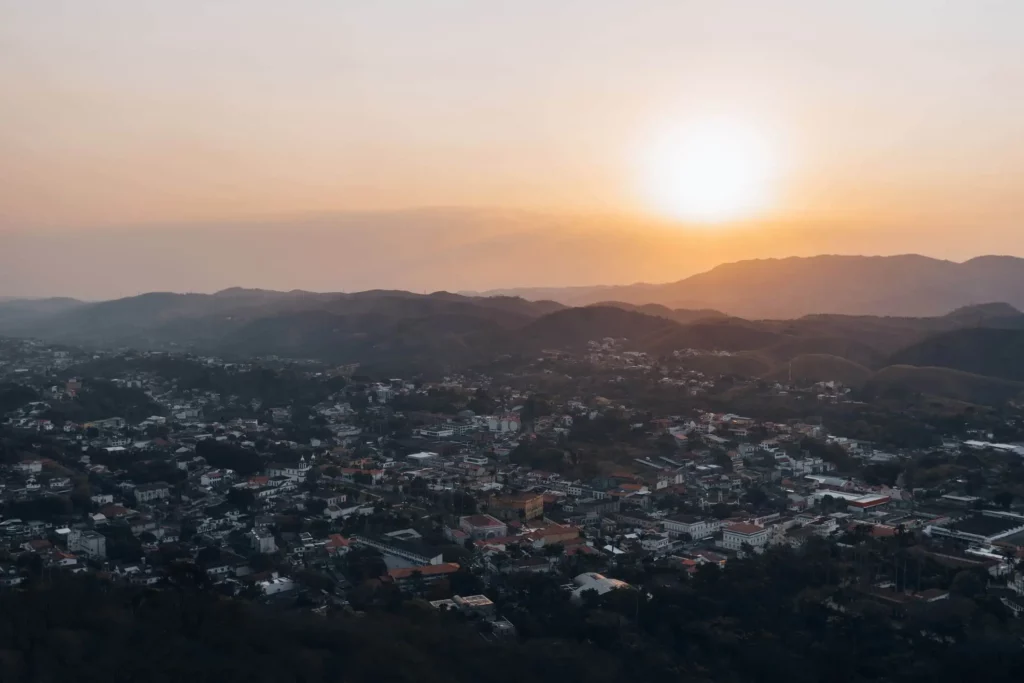 Imagem da vista aérea de Vassouras, no RJ, mostra colinas onduladas ao pôr do sol, com o sol baixo no céu lançando uma luz dourada quente sobre a paisagem e os edifícios