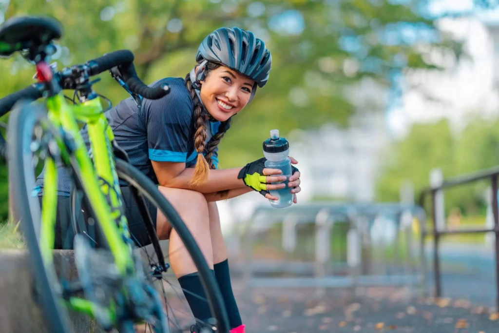 Imagem de uma ciclista sentada na calçada de um parque segurando uma garrafinha de água ao lado da bicicleta estacionada perto das árvores para ilustrar matéria sobre andar de bicicleta em São Paulo