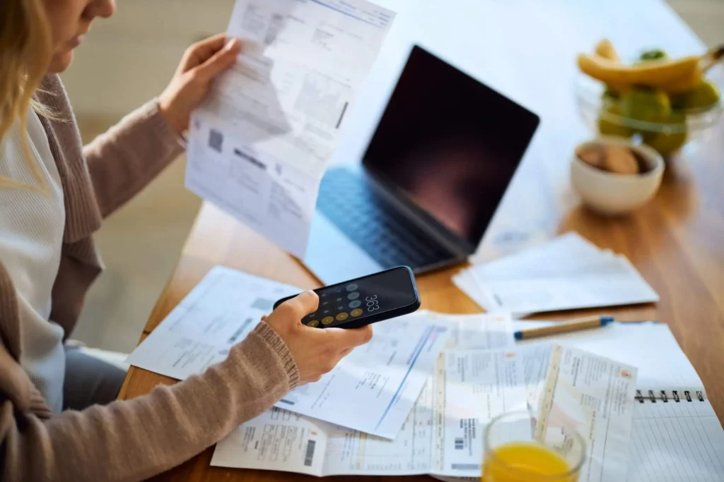 Imagem de uma mulher segurando um boleto em frente a um computador enquanto faz cálculos no celular para ilustrar matéria sobre como o IPTU é calculado