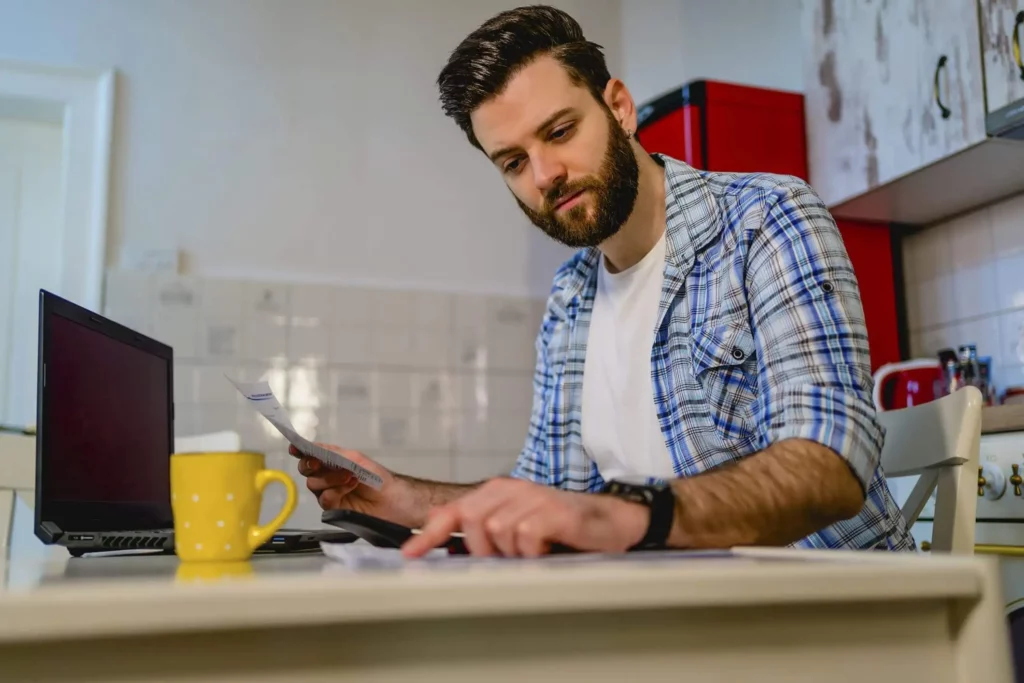 Imagem de um homem sentado ao redor de uma mesa onde estão um notebook, uma caneca de café e alguns papéis enquanto faz conta em uma calculadora para ilustrar matéria sobre como pagar IPTU atrasado em Santo André