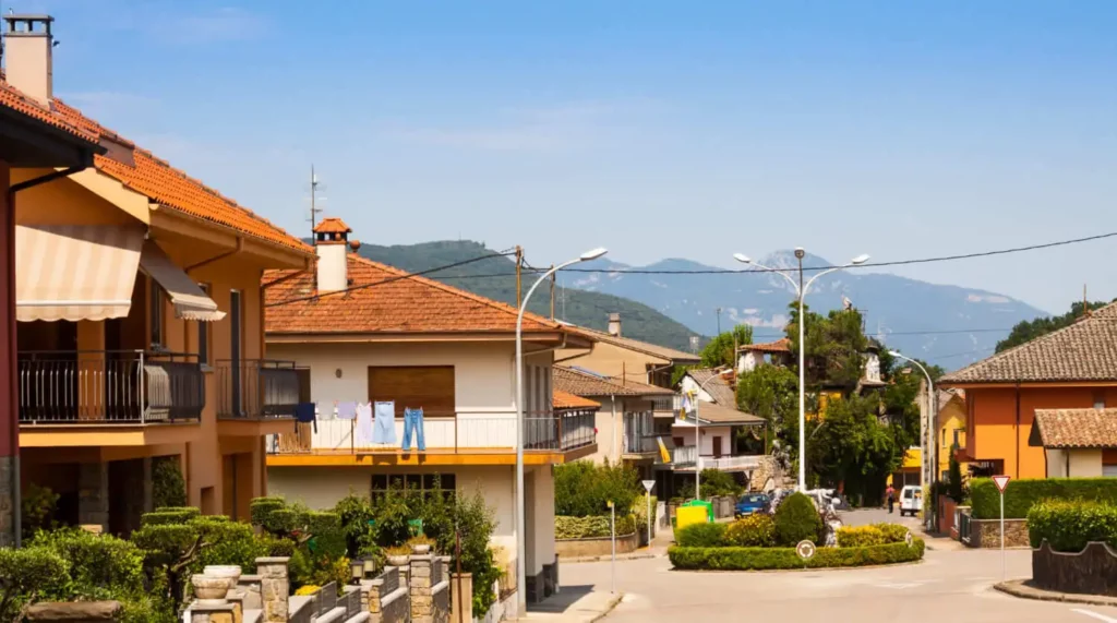 Imagem de um condomínio horizontal mostra casas com telhados de terracota, jardins, uma rua e montanhas ao fundo sob um céu azul claro.