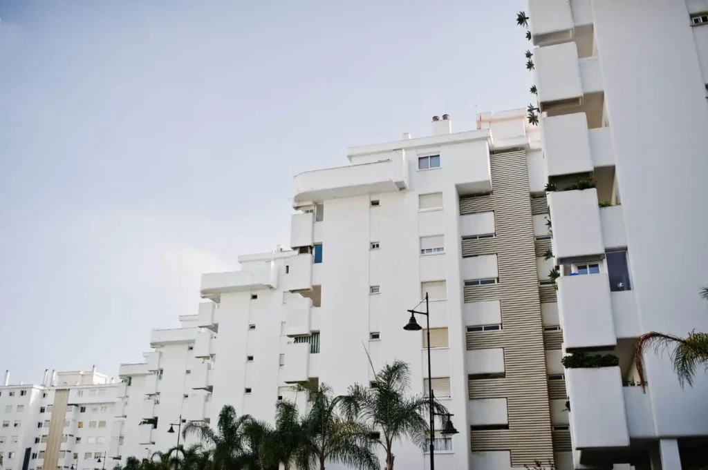 Imagem de vários prédios de apartamentos brancos e modernos com varandas retangulares se alinham sob um céu claro para ilustrar matéria sobre como é morar em um condomínio misto. Palmeiras e um poste de luz são visíveis em primeiro plano.