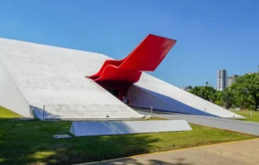 Fachada moderna do Auditório Ibirapuera, com sua arquitetura icônica em tons vermelhos, cercada por áreas verdes e sob um céu azul