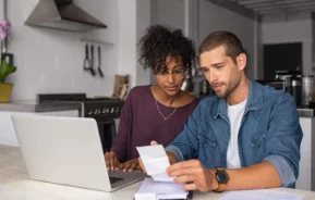 Imagem de um casal formado por um homem e uma mulher sentados ao redor de uma mesa que está na cozinha de casa enquanto fazem contas e olham alguns documentos que estão em cima da mesa ao lado de um notebook para ilustrar matéria sobre IPTU em Santo André