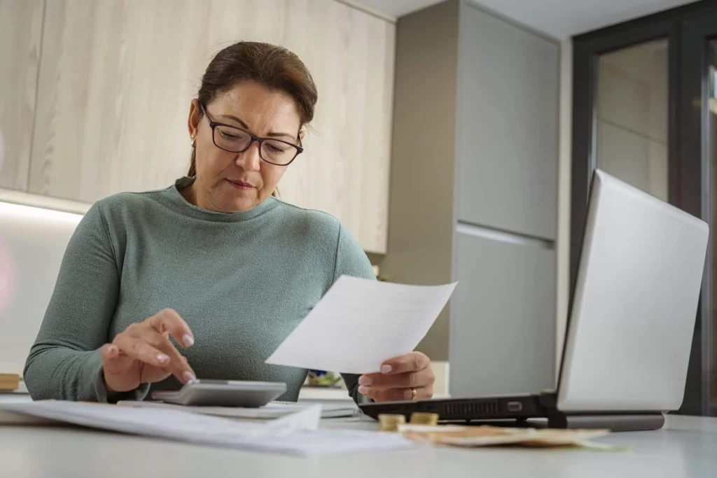 Imagem de uma mulher fazendo contas com o auxílio de uma calculadora que está em cima de uma mesa ao lado de alguns papéis e um notebook para ilustrar matéria sobre o IPTU na cidade de Santo André, no ABC Paulista
