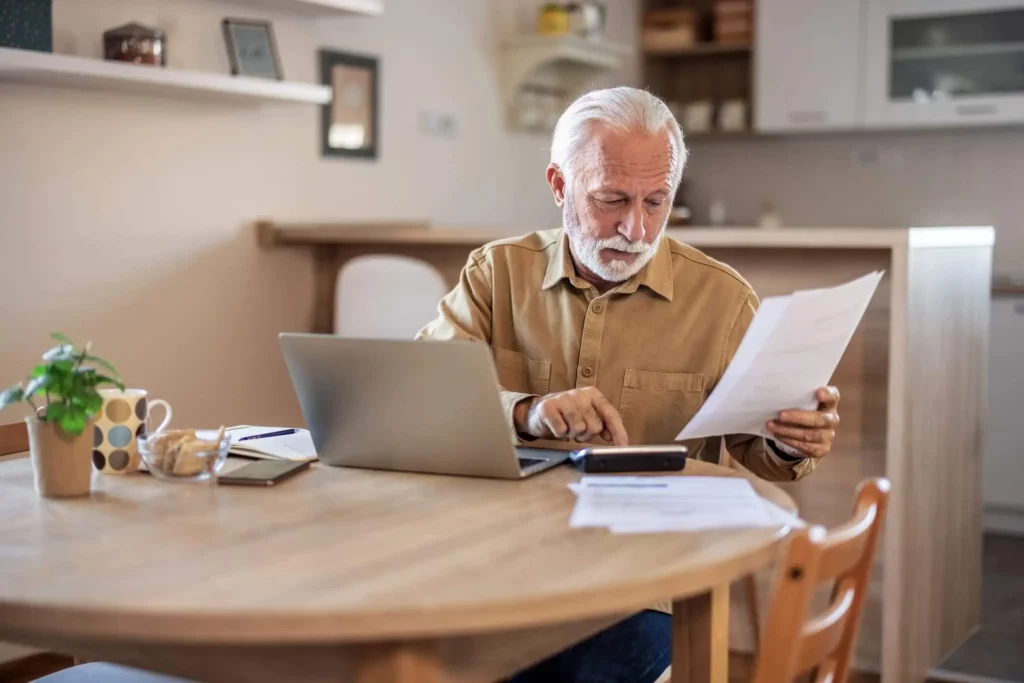 Imagem de um senhor sentado ao redor de uma mesa de madeira que está na cozinha olhando para as informações que estão em um papel enquanto faz contas em uma calculadora que está ao lado de um notebook para ilustrar matéria sobre a isenção de IPTU em Santo André