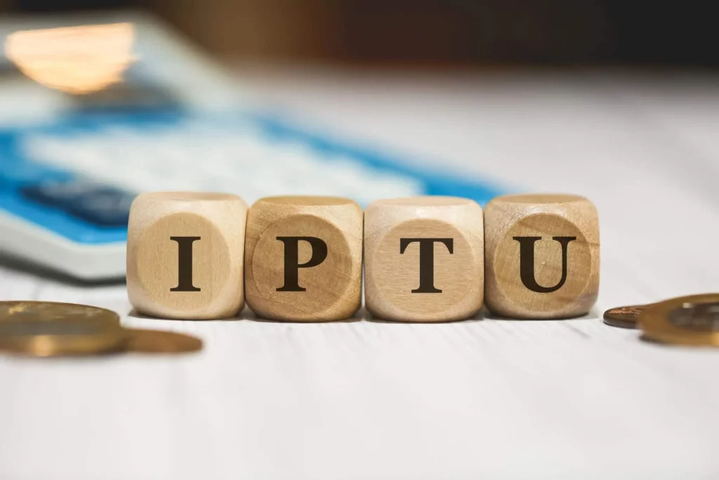 Imagem das siglas que formam a palavra IPTU escritas em quatro dados de madeira que estão ao lado de moedas e em frente a uma calculadora para ilustrar matéria sobre o que é taxa predial