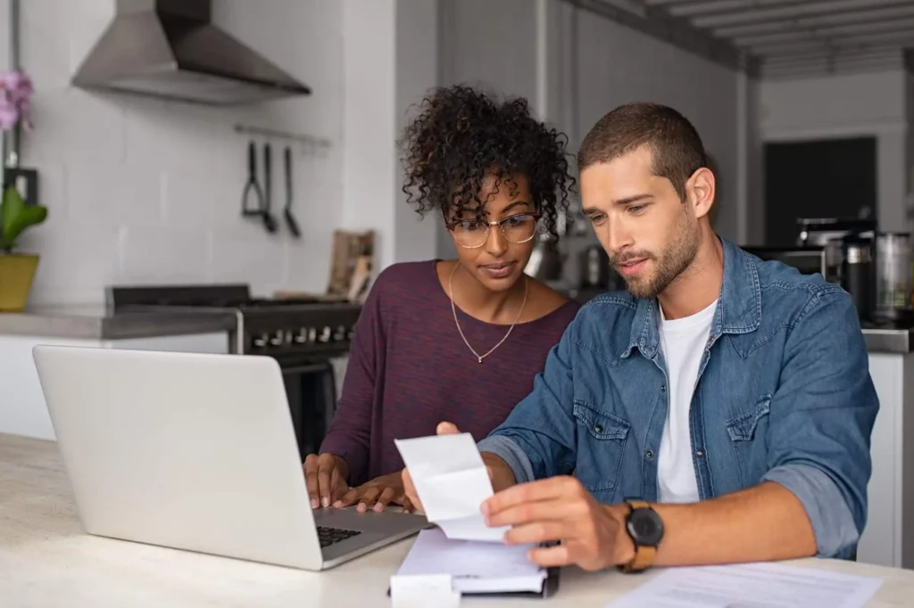 Imagem de um casal sentado de uma mesa em frente ao notebook analisando uma conta em papel na cozinha de casa para ilustrar matéria sobre o que é valor predial