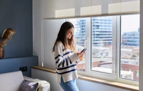 Imagem de uma mulher feliz usando o celular perto da janela de um apartamento para ilustrar matéria sobre qual a diferença entre studio e apartamento