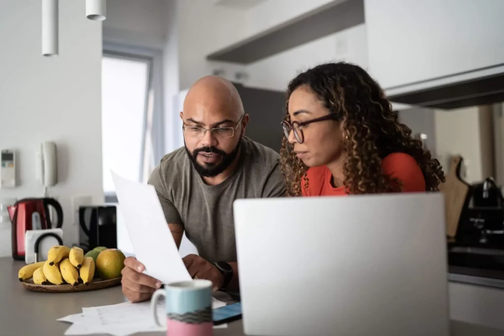 Imagem de um casal formado por um homem e uma mulher sentados ao redor da mesa da cozinha olhando as informações que estão em um papel ao lado de um notebook para ilustrar matéria sobre quem paga IPTU atrasado