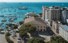 Foto mostra a fachada do Mercado Modelo em Salvador vista de cima, com a Baía de Todos os Santos ao fundo em um dia ensolarado e de céu azul (Foto: Márcio Filho - MTUR)