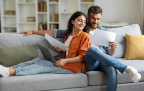 Imagem de um casal formado por um homem e uma mulher sentados confortavelmente no sofá da sala de uma casa enquanto olham as contas em alguns papéis para ilustrar matéria sobre o aumento do aluguel em 2025