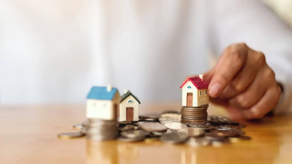 Imagem da mão de uma pessoa colocando algumas casas de madeira em tamanho miniatura em cima de algumas moedas que estão em uma mesa para ilustrar matéria sobre reajuste de aluguel em 2026.