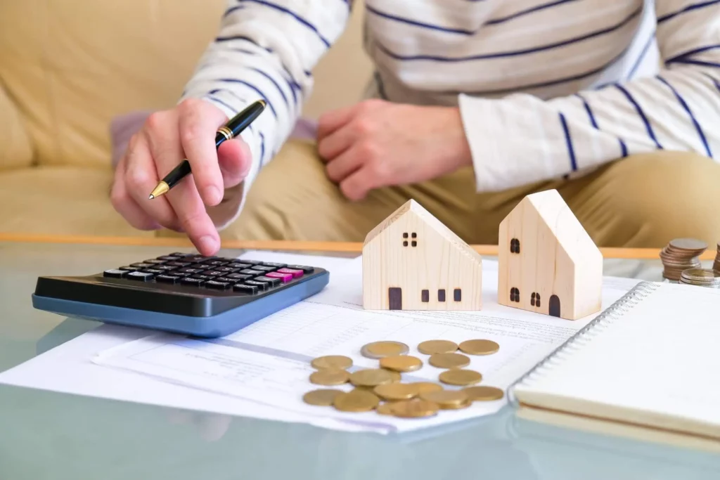 Imagem de uma pessoa fazendo contas em uma calculadora que está em cima de uma mesa de centro próxima a papéis, moedas e casas de madeira em tamanho miniatura para ilustrar matéria sobre o reajuste de aluguel baseado no IPCA