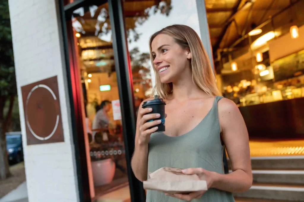 Imagem de uma mulher saindo de uma padaria com um copo de café para ilustrar matéria sobre o que é um bairro comercial
