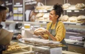 Imagem de uma atendente de padaria mostrando um pão artesanal para o cliente atrás do balcão para ilustrar matéria sobre as padarias em SP