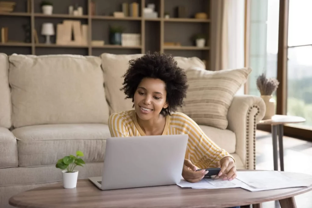 Imagem de uma mulher sentada no chão da sala enquanto olha o notebook que está em cima da mesa de centro para ilustrar matéria sobre o reajuste de aluguel 2026.