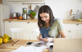 Imagem de uma jovem mulher sentada à mesa da cozinha, sorrindo, enquanto analisa um recibo para ilustrar matéria sobre reajuste de aluguel. Ela está cercada por papéis e um notebook em uma cozinha.