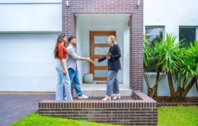 Imagem que ilustra matéria sobre fusão de imóveis mostra um casal em frente à uma casa dando as mãos para uma mulher