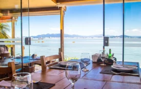 Foto que ilustra matéria sobre os melhores restaurantes em Florianópolis mostra o ambiente interno do Rosso Restaurante com uma vista para o mar através de uma grande janela de vidro (Foto: Divulgação)