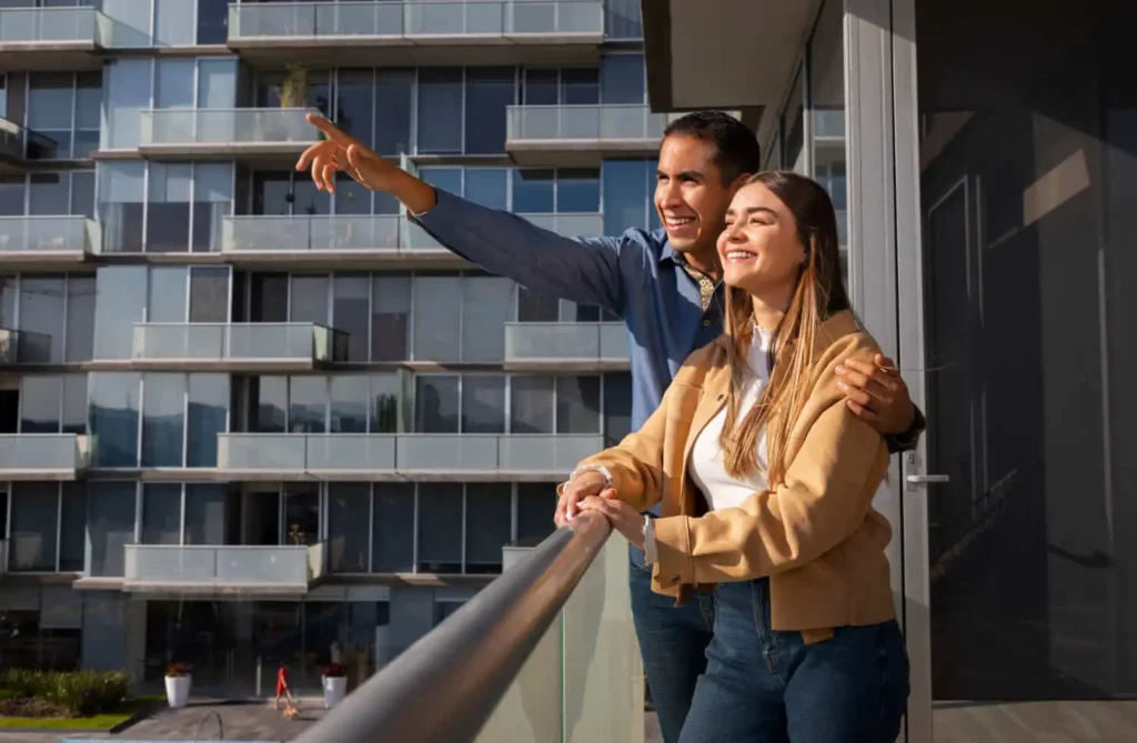 Imagem de um casal de jovens sorridentes na sacada de um prédio enquanto observam a vizinhança para ilustrar matéria sobre as características dos megacondomínios