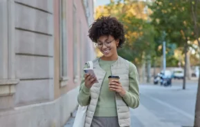 Imagem de uma mulher de cabelos cacheados, de óculos e com um colete bege sorri enquanto olha para o celular e segura uma xícara de café para ilustrar matéria sobre as cidades inteligentes do Brasil.