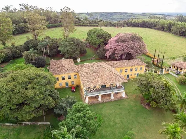 Vista aérea de uma grande casa de campo amarela com telhado de telhas, cercada por árvores e gramados verdes que lembram os parques de Campinas, com campos abertos e colinas ao fundo.