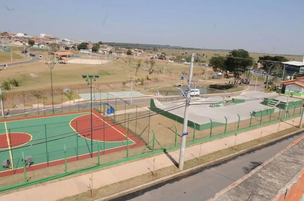 Vista aérea de um complexo esportivo ao ar livre no bairro Parques, em Campinas, que conta com uma quadra de basquete, um parque de skate, gramados e uma pista de caminhada. A área é cercada e fica próxima a uma estrada, com prédios residenciais e terrenos ao fundo.